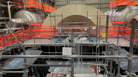 Metal scaffolding and bright orange netting cover the room, while the light-brown metal arch of the partially dissassembled turbine cover sits inside the Palisades nuclear plant.
