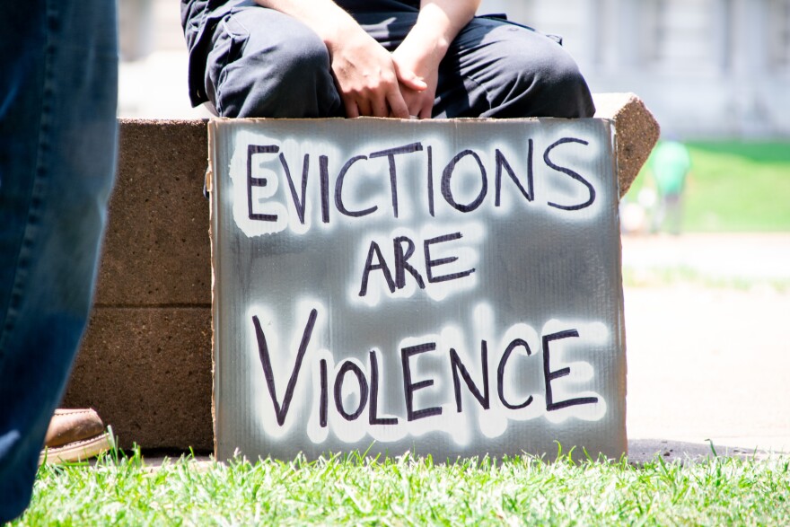 A sign reading "Evictions Are Violence" rests against the legs of a person sitting on a stone bench.