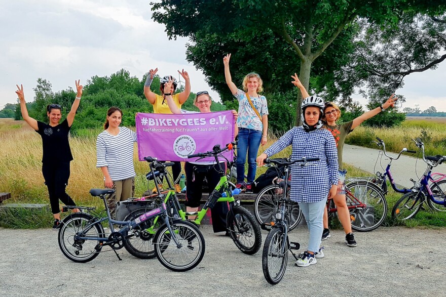 Volunteers and trainees with the group Bikeygees at a park in Berlin in July. The organization teaches refugee women in Germany how to ride bikes. Trainee Shapol Bakir-Rasoul, a refugee from Iraq, holds up a Bikeygees sign with founder Annette Krüger, right. Behind them in yellow is volunteer Shaha Khalef, a refugee from Iraq.