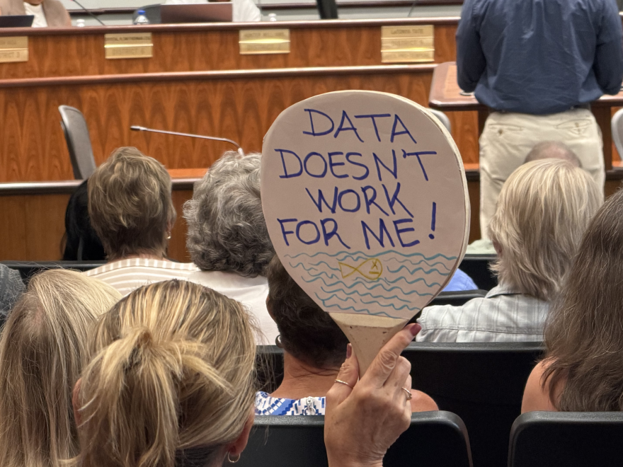 A protester holds up a sign in a Birmingham City Council meeting protesting data center development that says, "DATA DOESN'T WORK FOR ME." It depicts a dead fish in a body of water.