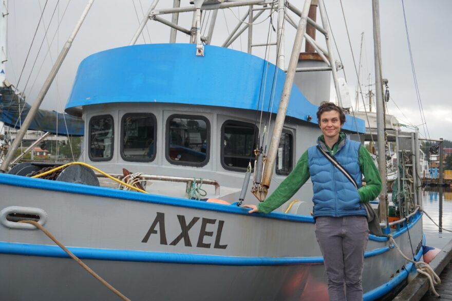 A white woman in a blue best poses next to a light blue boat with teh name "Axel"