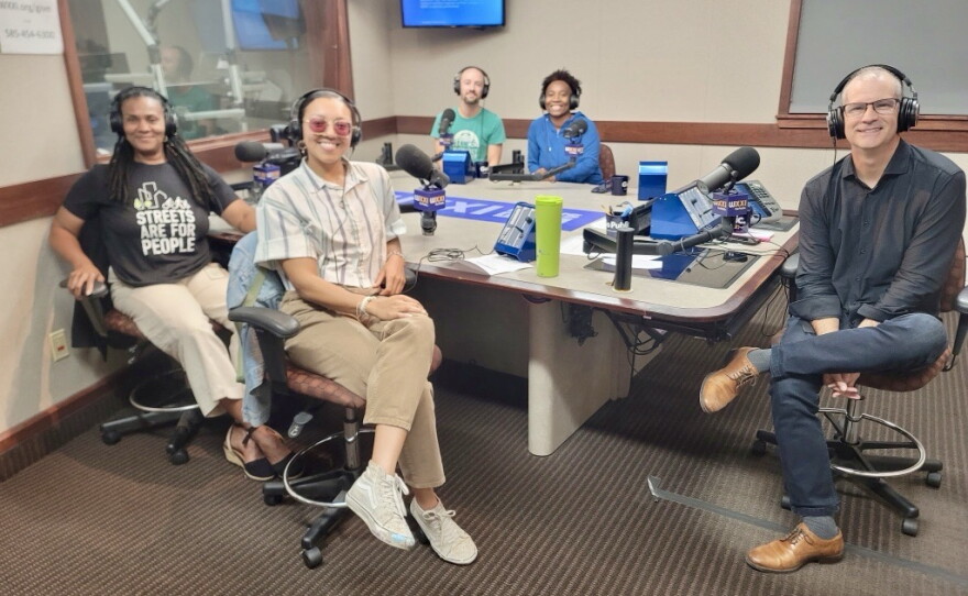 Five smiling people wearing headphones sit around a table in a radio talk studio: A woman front left has long black braids and is wearing white pants, black shoes, and a black graphic t-shirt with white text that reads "STREETS ARE FOR PEOPLE;" a woman front center has her dark hair tied back and is wearing sunglasses, a blue and white collared button-down shirt, tan pants, and white sneakers; a man back left is bald and is wearing a green graphic t-shirt; a woman back center has short black hair and is wearing a blue top; a man at right has very short dark hair and is wearing a black button-down shirt, jeans, and brown sneakers