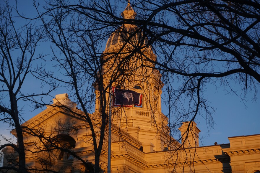 
The sun sets on a government building, seen behind winter bare trees and a Wyoming flag.