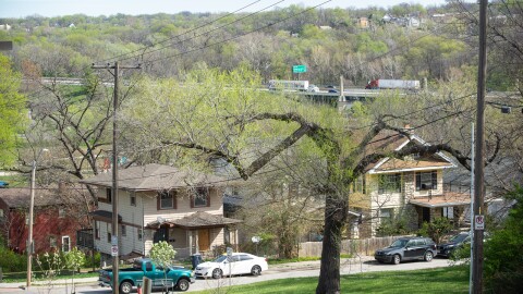 A pair of two-story houses sit in the foreground with cars parked in the street. In far background, a highway can be seen with traffic moving along it.