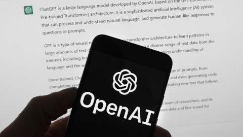 The OpenAI logo is seen on a mobile phone in front of a computer screen which displays output from ChatGPT.