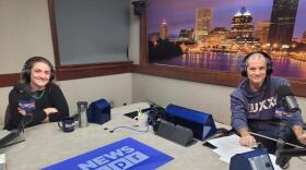 Two people wearing headphones sit at a table in a radio talk studio: a woman at left has long brown hair and is wearing a black sweater with a green beaded necklace; a man at right has short dark hair and is wearing a blue hooded sweatshirt with white lettering.