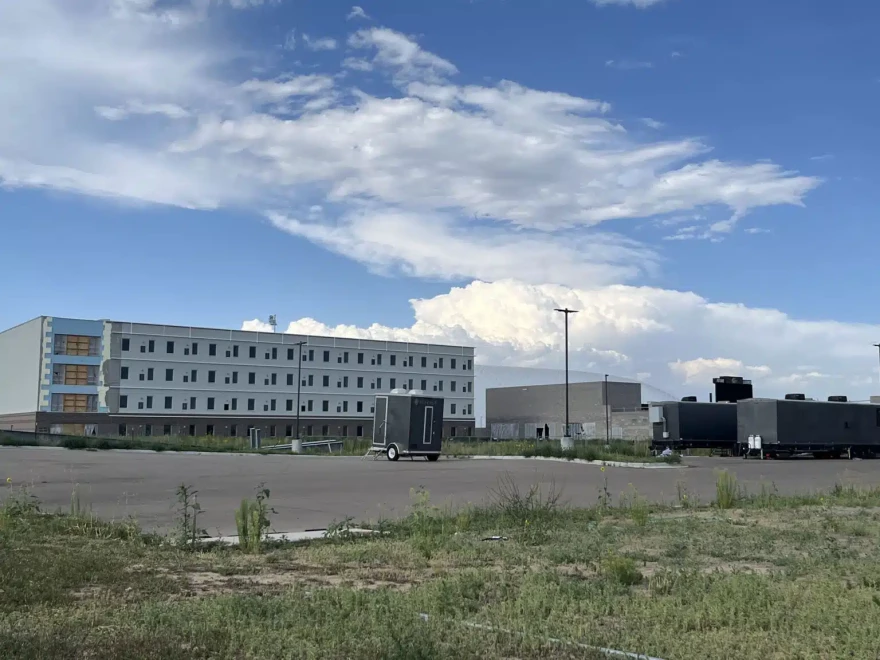 A long white building sits in a field of unkempt, patchy weeds. In front of the building, a small gray trailer has been left in an otherwise empty parking lot.