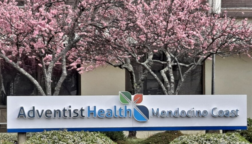 A sign reading "Adventist Health Mendocino Coast" underneath trees with pink blossoms.