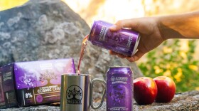 Cans of cider and apples are positioned on a rock outdoors, as a hand pours some into a silver mug. 