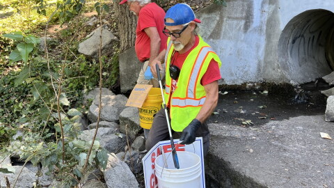 Two middle-aged men in red shirts use trash grabbers to pick up trash in a concrete culvert. 