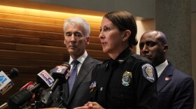 A woman in a police uniform stands at a podium. Two men stand behind her. 