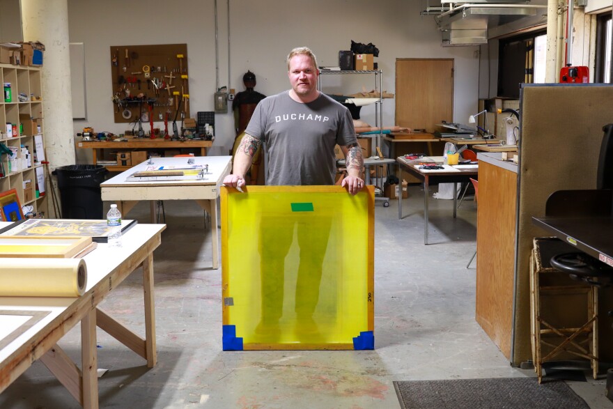  Osmon stands in the screen printing station at Factory Two. Two long tables are arranged to his left. In the background, there are shelves full of supplies, and tools mounted on the wall. 