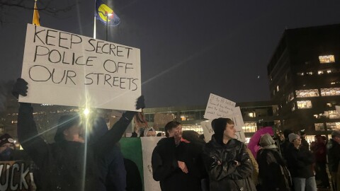 Standing in darkness, a protester holds a sign above their head, which reads "KEEP SECRET POLICE OFF OUR STREETS" Other protesters stand alongside them, some holding signs in the distance.