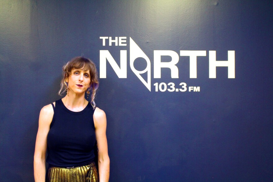 Mara Lovejoy posing in front of a blue wall that reads "The North 103.3 FM" in white letters. The "o" in north is designed to look like a compass.