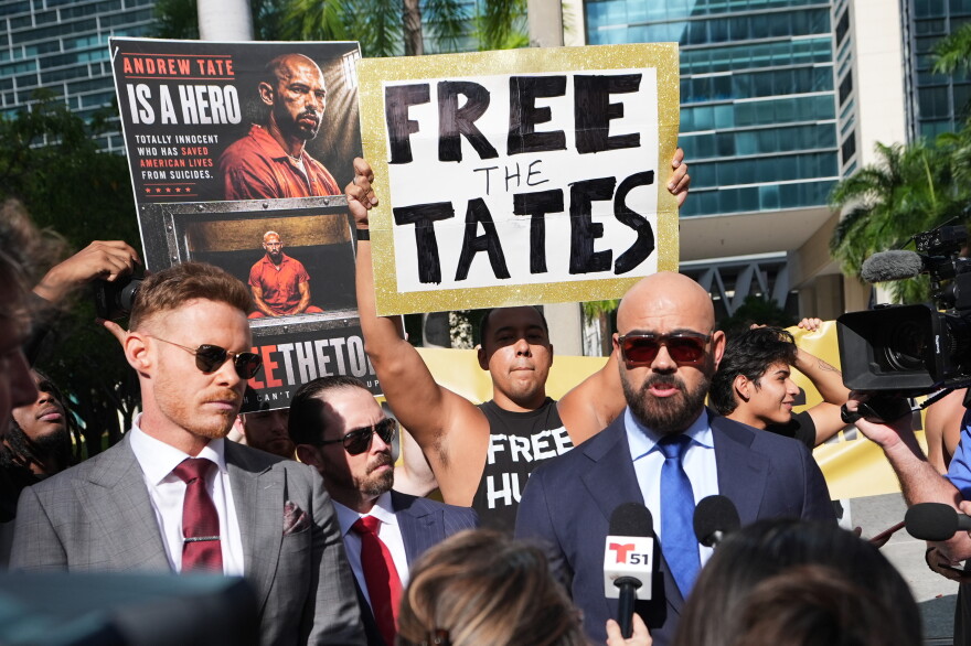 Two suited men speak on microphones while men behind them hold up signs. 