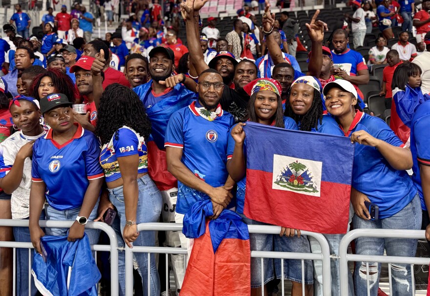 A large crowd in a stadium wearing blue and red jerseys, with two people holding a Haitian flag. 