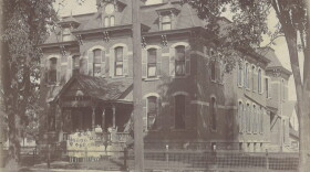  A black and white photo from about 1910 of the brick Second Empire-style building that was the Kalamazoo Children's Home for girls. The building had a distinctive mansard roof. Several residents are standing on the front porch in white dresses.  