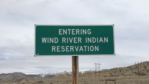 A green sign that reads "Entering Wind River Indian Reservation" stands in front of a background of grasslands, fences, telephone lines and snow-capped peaks. 