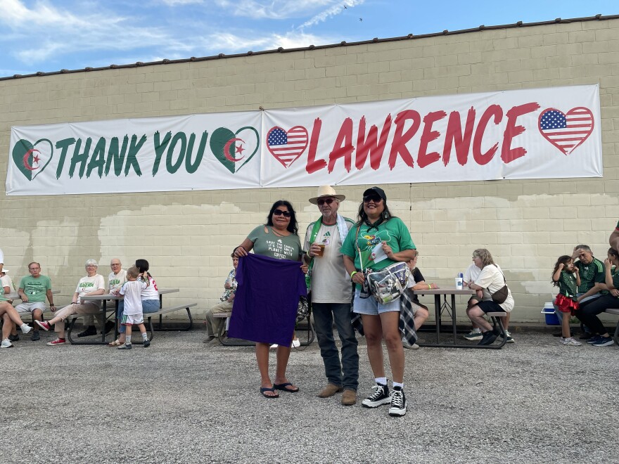 (From left) Laurie LaCour, Nez Perce Tribe and Confederated Tribes and Bands of the Yakama Nation; Stan Herd, Lawrence earthwork artist who created the Algerian flag on the Lied Center lawn of KU; and Jennifer Lewis, White Mountain Apache Tribe, pose in front of the “Thank You Lawrence” sign created and donated by Algeria supporters attending the team’s final block game with Austria at Arrowhead Stadium.