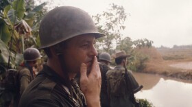 A photo of Bob Schieffer, a white man wearing a green army helmet and fatigues, smokes the stub of a cigarette while staring out over a brown river. Behind him are wide-leafed tropical trees.