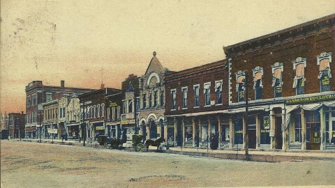 A colorized postcard showing a street view of the East Side Public Square in Bedford, Indiana during the early 1900s, featuring multi-story brick storefronts, horse-drawn carriages along the unpaved street, and text at the bottom reading "East Side Public Square, Bedford, Ind."