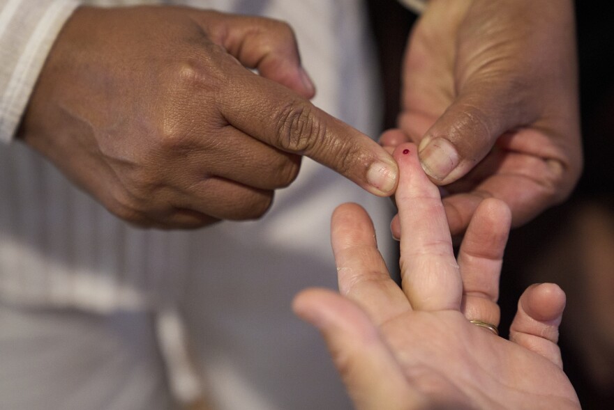 A small drop of blood is squeezed out of the tip of a finger by another set of hands.