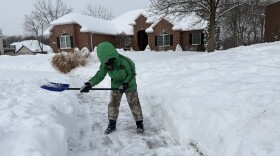 A child is surrounded by snow. He's wearing a bright green winter coat, camo snow pants, boots and gloves is shoveling snow off the sidewalk.