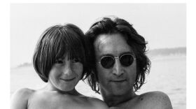 Black-and-white photo of a man wearing round sunglasses posing closely with a child, with ocean water in the background.