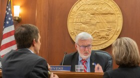 A white bearded man sits at a desk in front of a gold seal while two people talk to him.