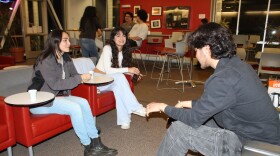 In the foreground, two people with long dark hair sit on red chairs across from another person with shorter dark hair. In the background, three other college students chat around a circular table.
