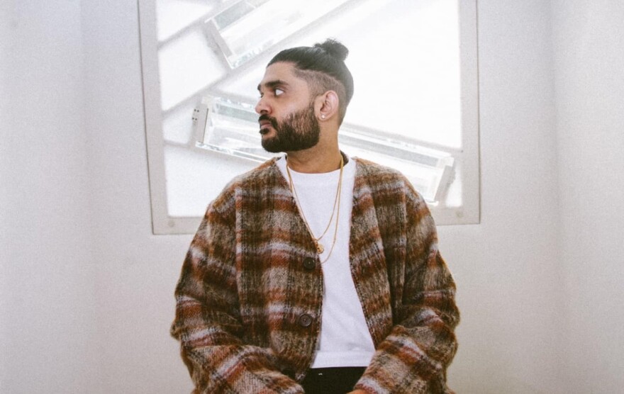 A man wearing an unbuttoned wool shirt over a white t-shirt looks to the left while sitting in a room with light-colored walls and an illuminated piece of art directly behind him.