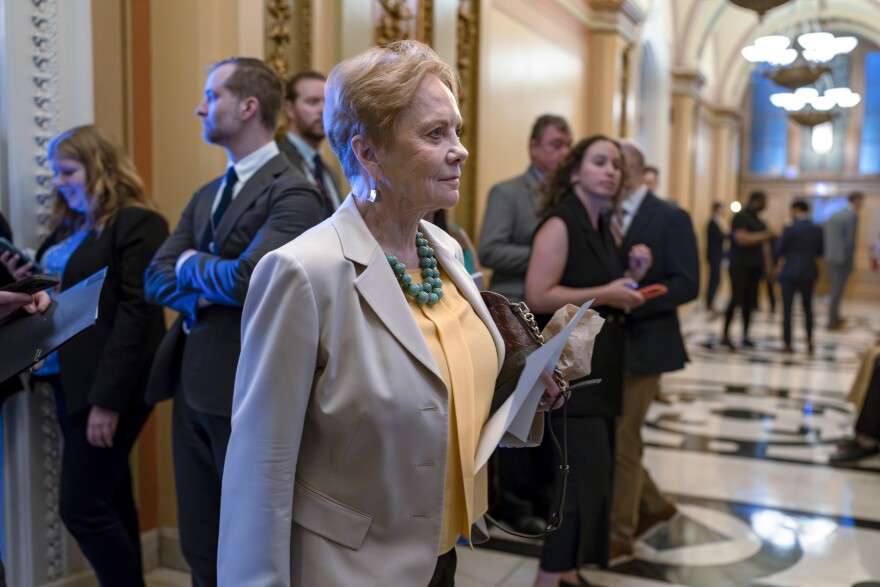 Kay Granger, an older woman with short reddish blonde hair wearing a gray suit, strides across a marble floor in a crowded Capitol building hallway, carrying a purse and some papers in one hand.