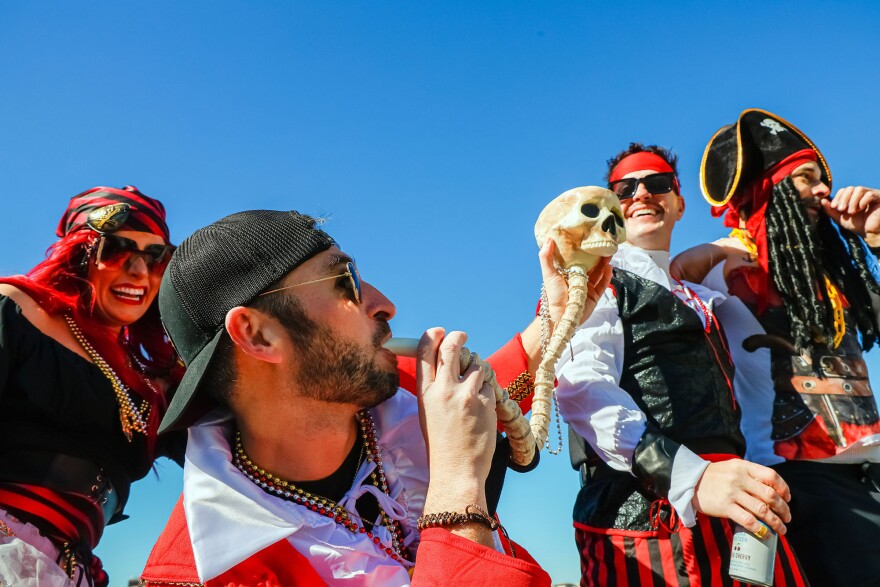 A man with a backwards baseball cap blowing into a horn with a skull at the end with people in pirate outfits smiling behind him
