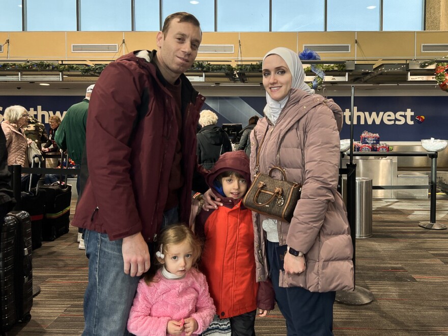 A man with a burgundy coat stands next to a small child in a pink sweater and a boy in an orange coat. A woman with a pink puffer coat stands on the right.