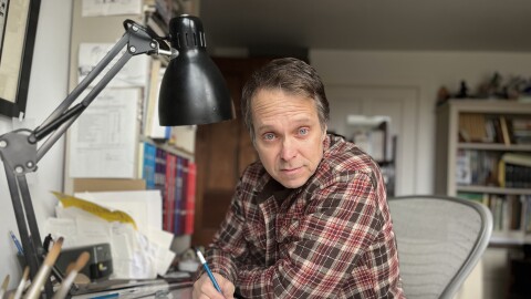 A man with short salt and pepper hair is wearing a plaid button-down shirt and drawing on a pad while sitting at a desk in an office.