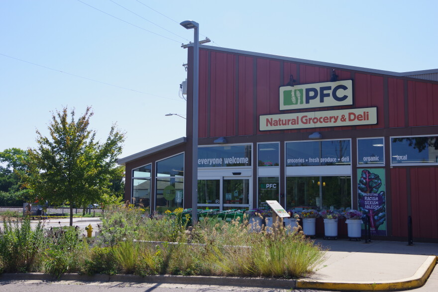 a red building with a slanted roof sits in front of thin flowers, weeds and other plants on a concrete walking path. The sign for the store reads "PFC Natural Grocery & Deli."