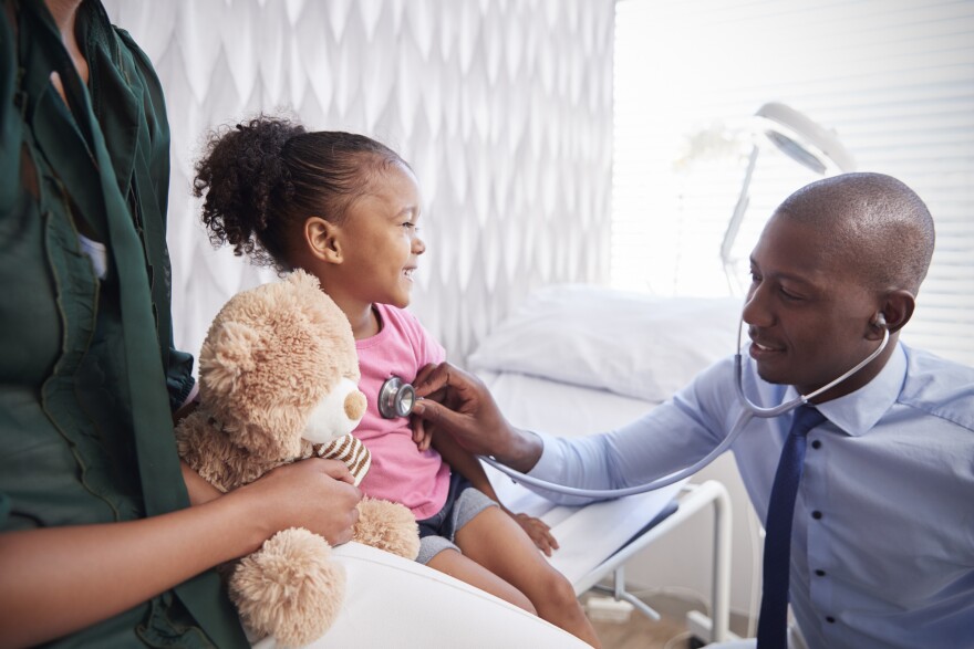 A doctor holds a stethoscope up to a young child's chest in a white medical exam room. The child is being held by an adult, who is also holding a teddy bear.