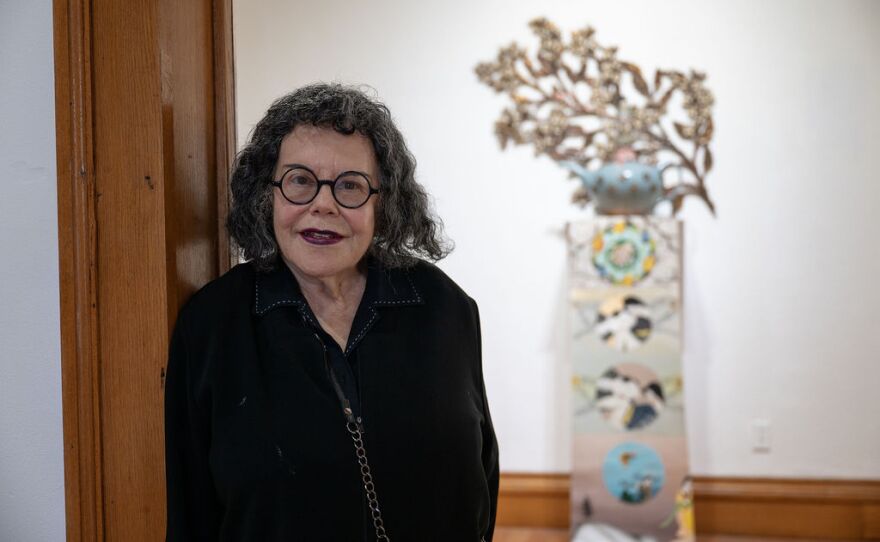 A woman wearing glasses and a black outfit standing in a doorway with a teapot sculpture in the background.