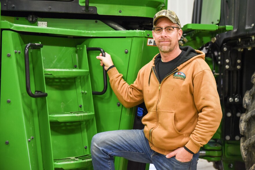 Farmer Mark Metz, in jeans and ballcap, stands next to his green farm equipment.