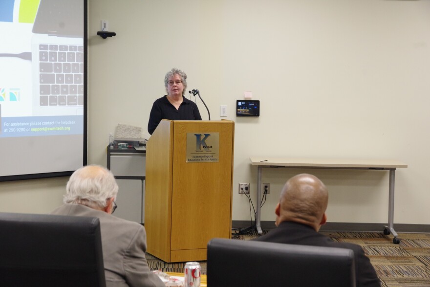 A woman in a black blouse stands at a light brown podium, part of a projector can be seen at the left side of the image. Two mean in the foreground sit in chairs, looking towards her as she speaks. 