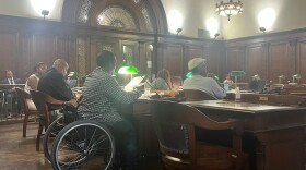 Five Albany Community Police Review Board members, pictured from behind, sit at wooden desks with green desk lamps. One member, who is closest to the picture and in a wheelchair, is reading from a sheet of paper.