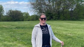 A woman stands in the middle of a grass field and trees behind her.