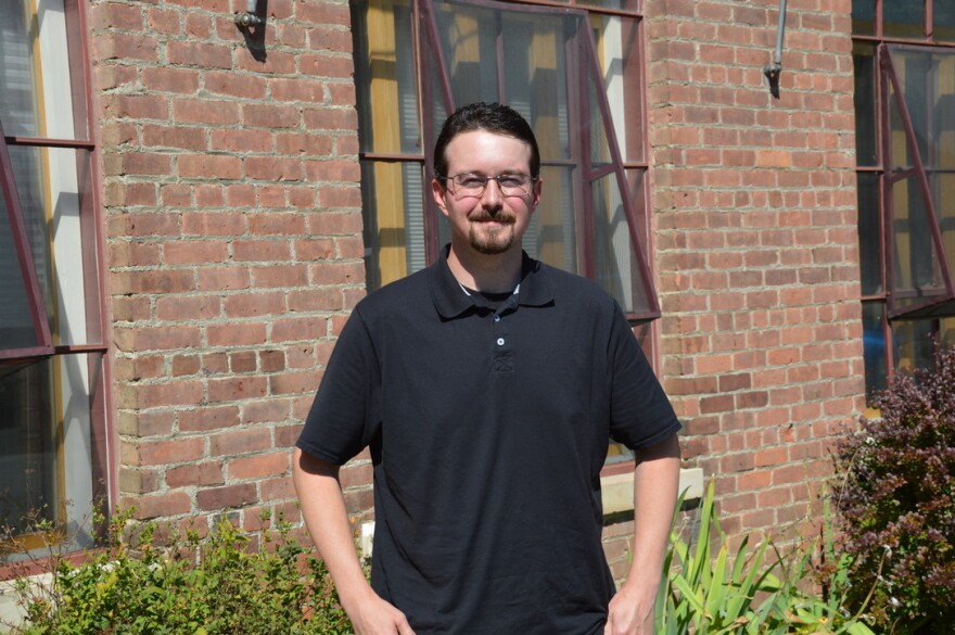 A young man in a black T-shirt stands in front of the red brick office building he works in.