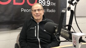 A man wearing glasses and a black Columbia jacket sits smiling at a radio station desk with a WCBU 89.9 FM microphone and banner visible in the background.