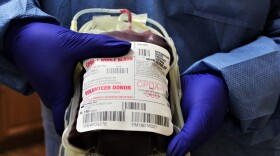 Blue-gloved hands hold a bag of blood that says 'volunteer donor.'