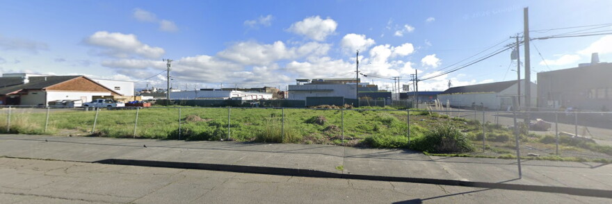 An empty site is covered with green grass and lined by chainlink fence. It's surrounded by white industrial buildings. 