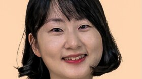 A smiling woman with short black hair and side-swept bangs poses against a soft peach background. She has a warm expression and is centered in the frame.