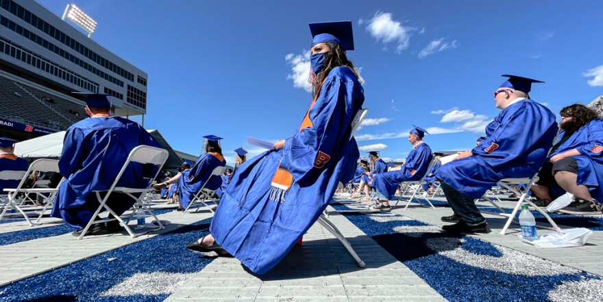With only a few wispy clouds in the sky, 2021 Boise State University graduates sit on plastic folding chairs on the blue turf of the school's football field. The focus is on one sole female graduate as she sits forward while everyone else looks behind her. The sky-box, for VIP fans, lingers in the background. 