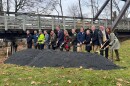 Gov. Glenn Youngkin (center), with 12 other federal, state and local officials, gather around a mound of grey crushed gravel, the same material used in the Virginia Creeper Trail. They hold shiny shovels, aimed at the mound. Behind them is a wooden bridge from the trail. 