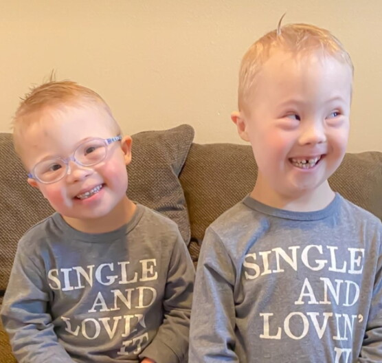 Two young boys in gray sweatshirts smile while sitting on a brown couch.